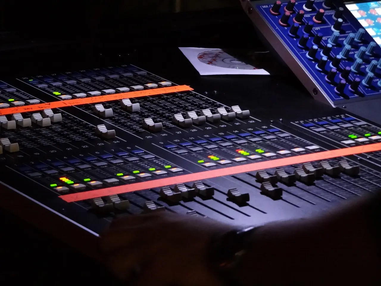 Detalle de una consola de mezcla digital con faders y luces indicadoras en un cuarto oscuro.