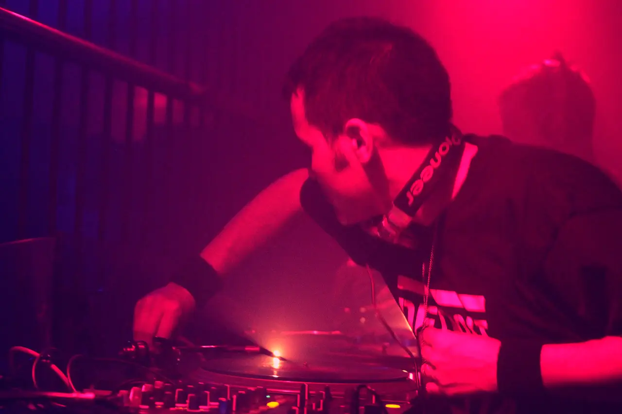 DJ inclinado sobre la mesa de mezcla con audífonos, bajo luz magenta y azul.
