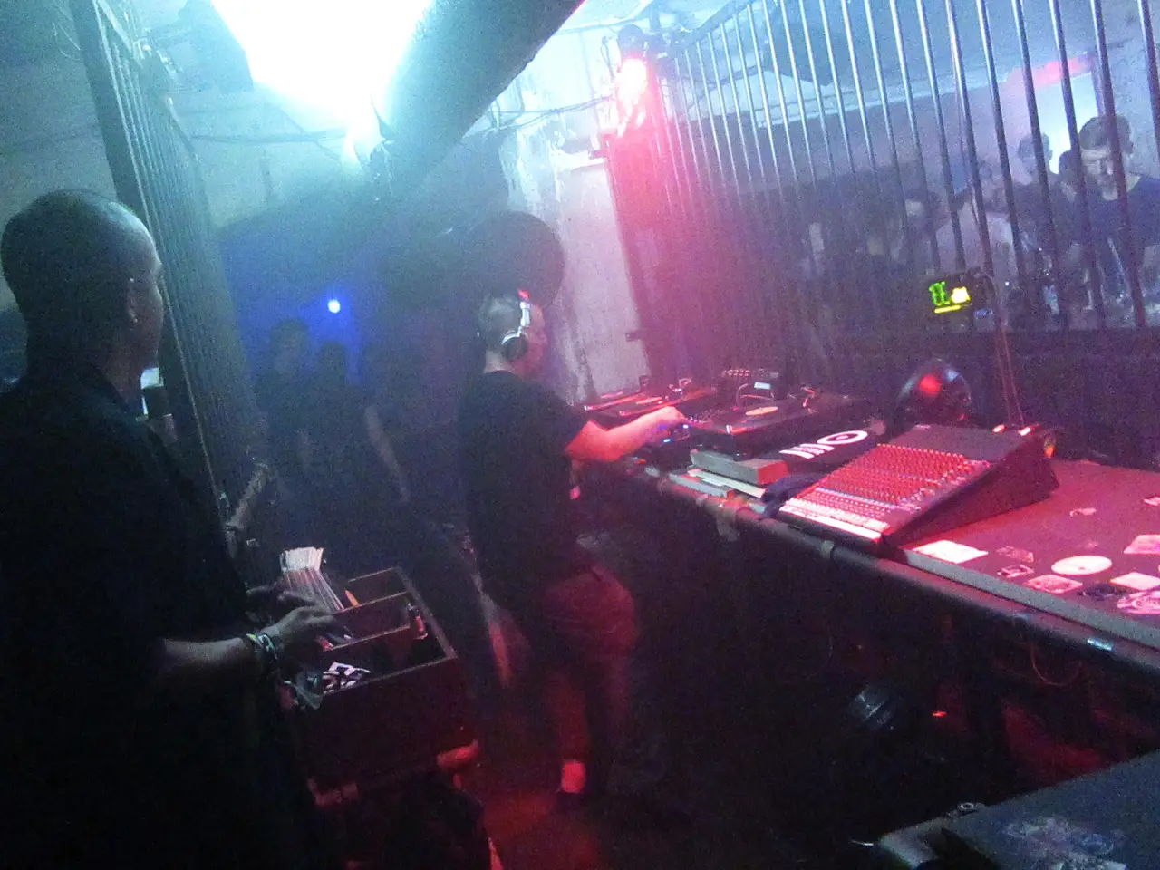 Cabina de DJ vista de lado en un club, con humo, luz roja y azul y público tras una reja.