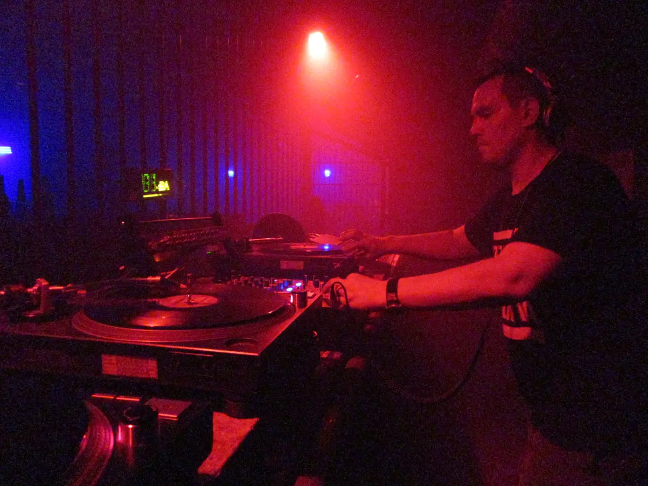 DJ de perfil con audífonos mezclando en dos platos, con luz roja y neón azul al fondo.