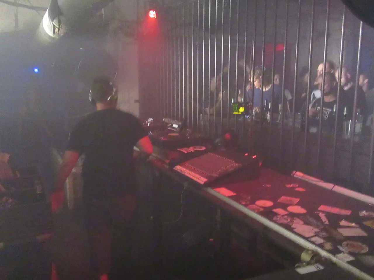 DJ de espaldas frente a la mezcladora, con humo y público tras la reja en un club oscuro.