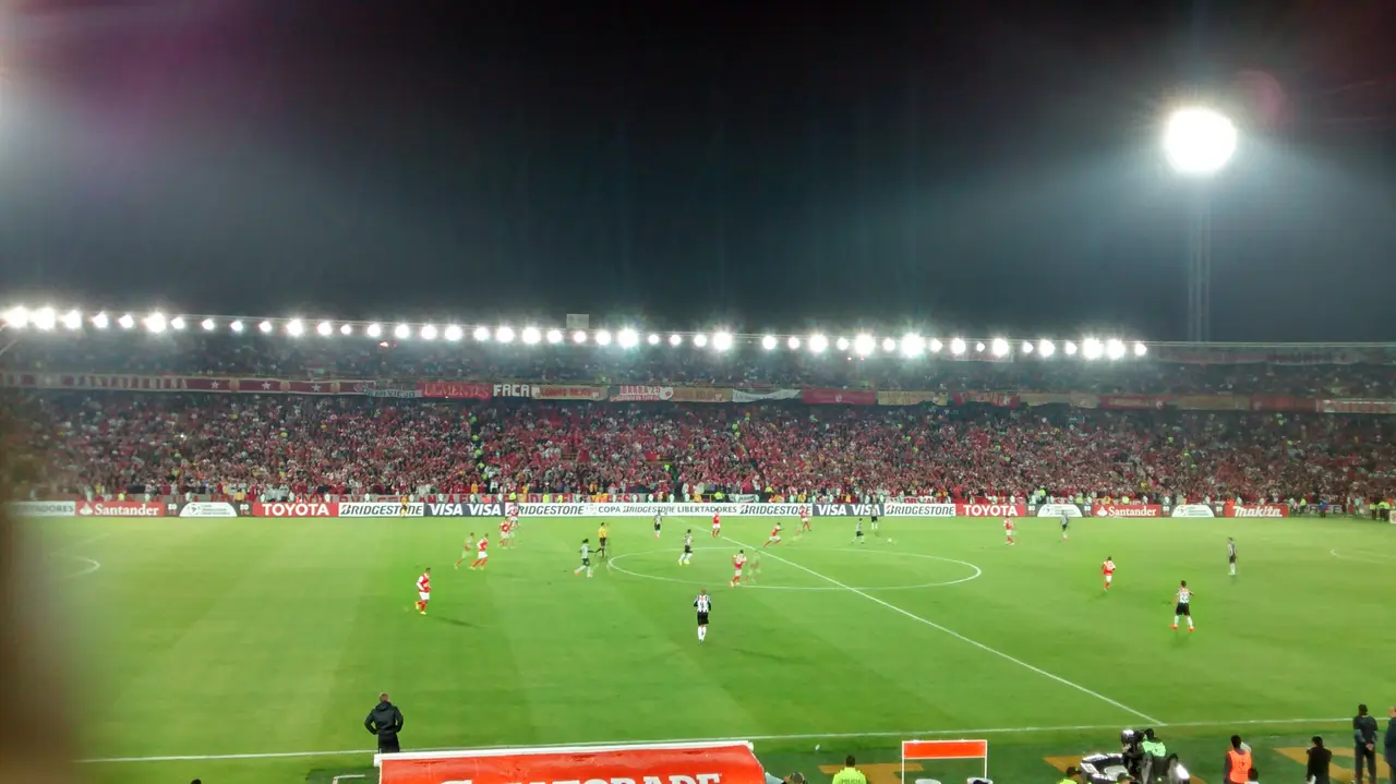 Vista general de la cancha iluminada de noche, con jugadores en movimiento y tribuna roja detrás.