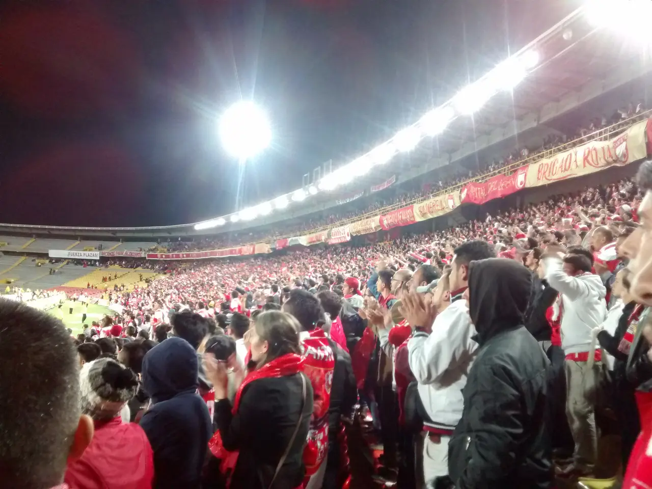 Hinchada con buzos y bufandas en la tribuna bajo los reflectores, de noche.