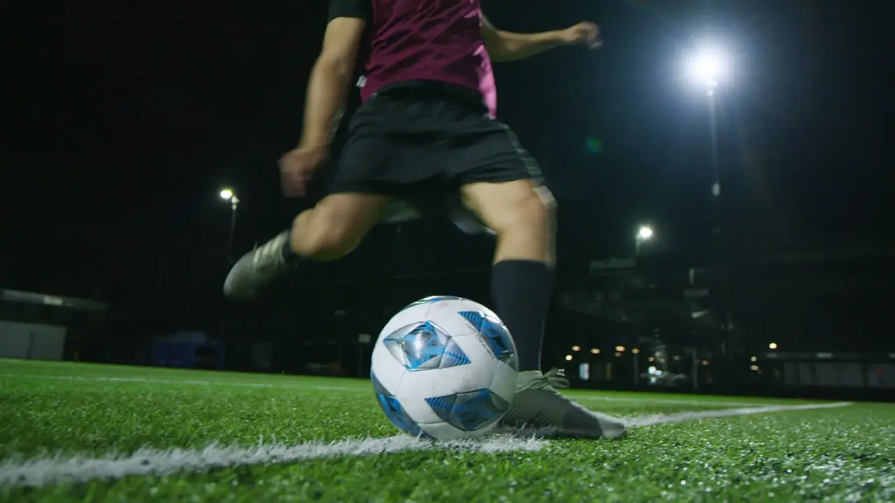 Plano bajo de un jugador a punto de patear el balón en una cancha sintética de noche, con reflectores al fondo.