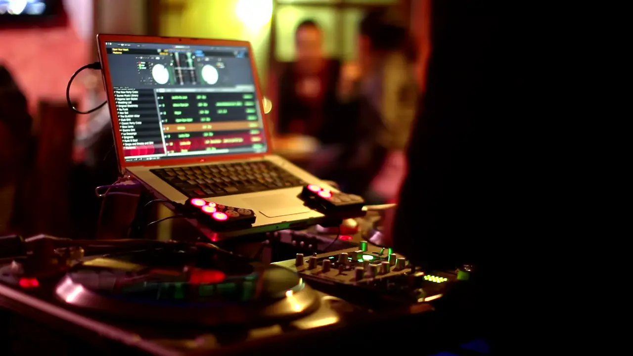 Laptop con software de DJ sobre una mesa con platos de vinilo y luces cálidas de club al fondo.
