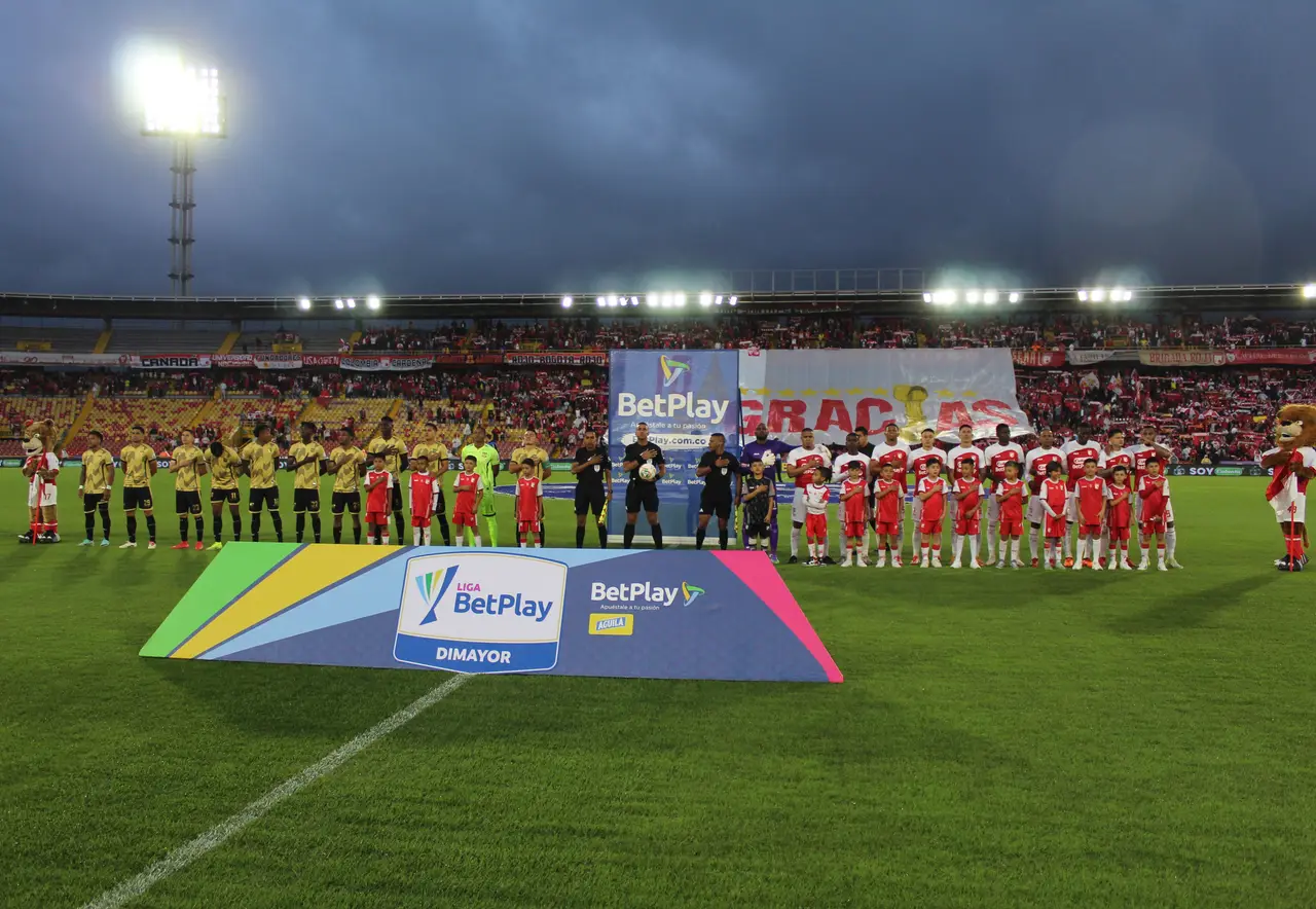 Dos equipos formados sobre el césped antes de un partido de noche, con la tribuna iluminada al fondo.